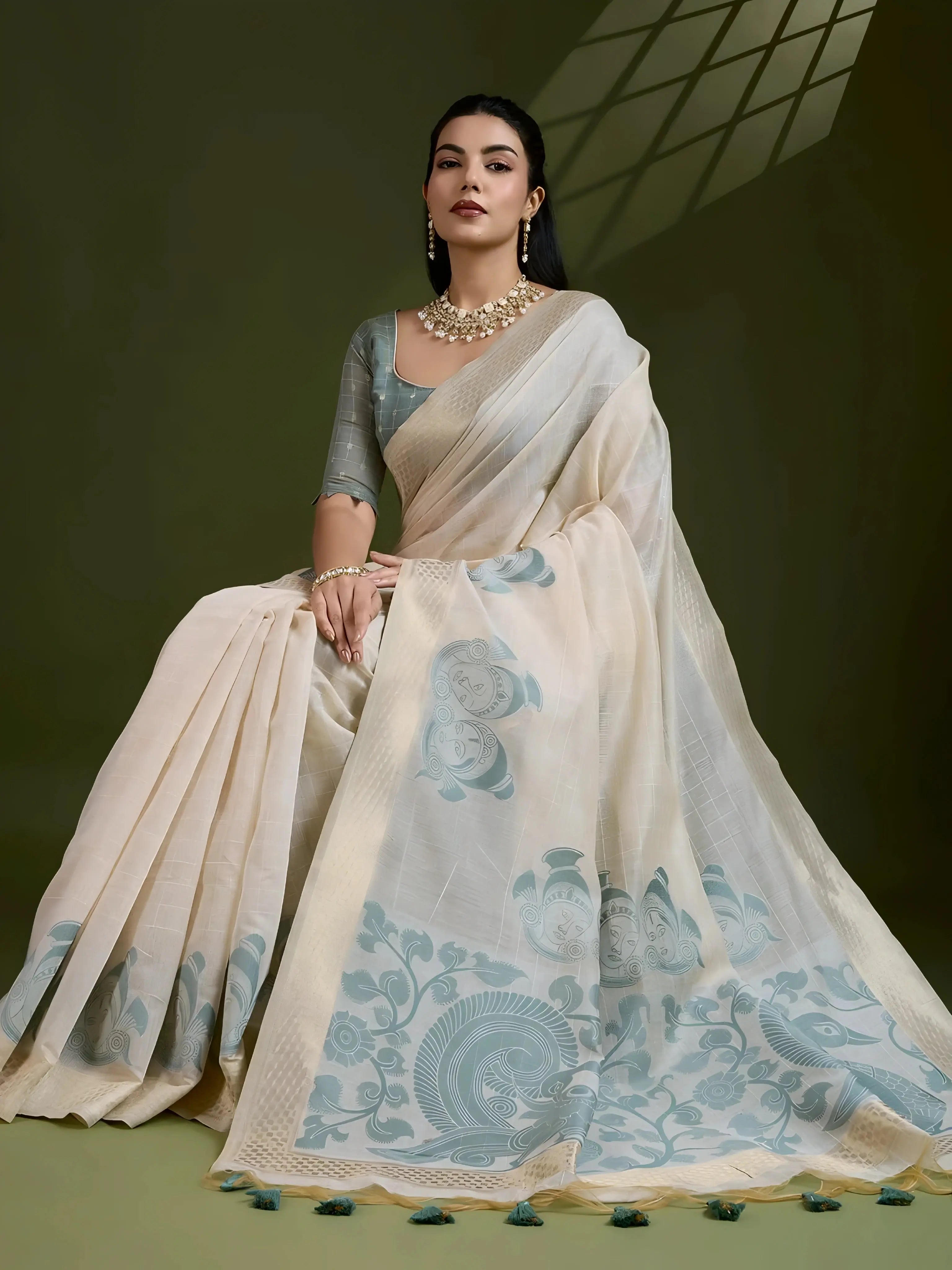 A woman models an elegant Beige Soft Muga Cotton Saree. The saree features large Kalamkari-style prints of peacocks and trees in a contrasting Dusty Sea Green on the pallu. She wears a sage green short-sleeve blouse and accessorizes with a gold and pearl Kundan choker necklace, posing indoors.